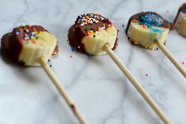 Banana Pops are as easy as putting banana slices on wooden skewers, covering in melted chocolate, and sprinkling sugar or other yummy toppings! Great recipe to make with the kids this summer. 