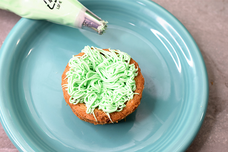 Cookie cup filled with green frosting that looks like grass sitting on a blue plate with a piping bag next to it.