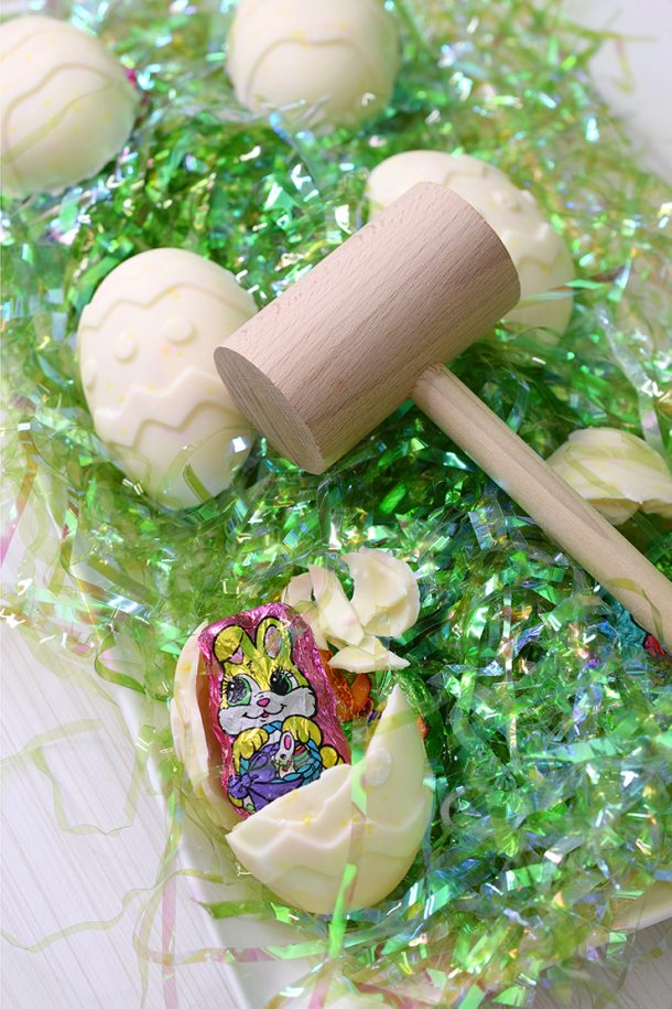 Broken chocolate piñata Easter Egg setting on platter next to wooden mallet and unbroken eggs on green Easter grass.