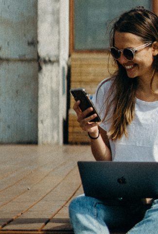 Woman holding a laptop and looking at a phone.