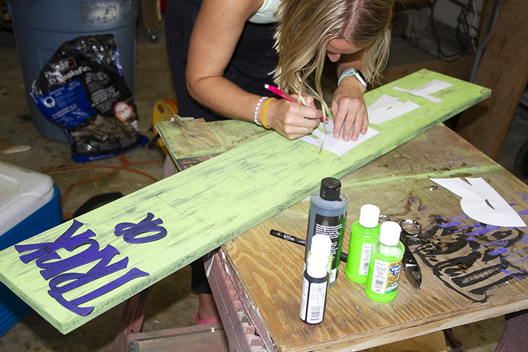 Blond woman painting letters on a large green, wooden sign.