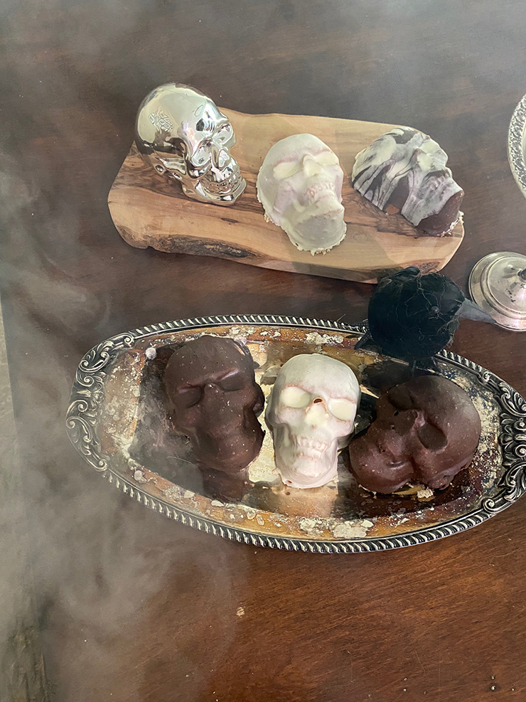 Mini cakes in the shape of skulls with brown and white frosting, sitting on a table with smoke all around them.