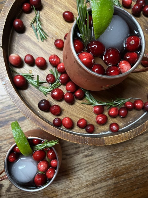 Yule Mule drink in a copper mug with a large, round ice cube, sitting on a wooden platter surrounded by cranberries.