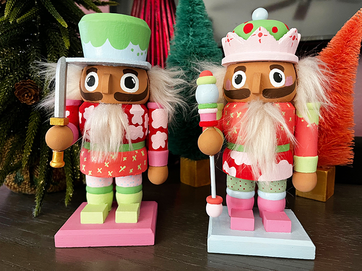 Two painted nutcrackers, in non-traditional pastel colors, in front of brightly colored christmas trees.