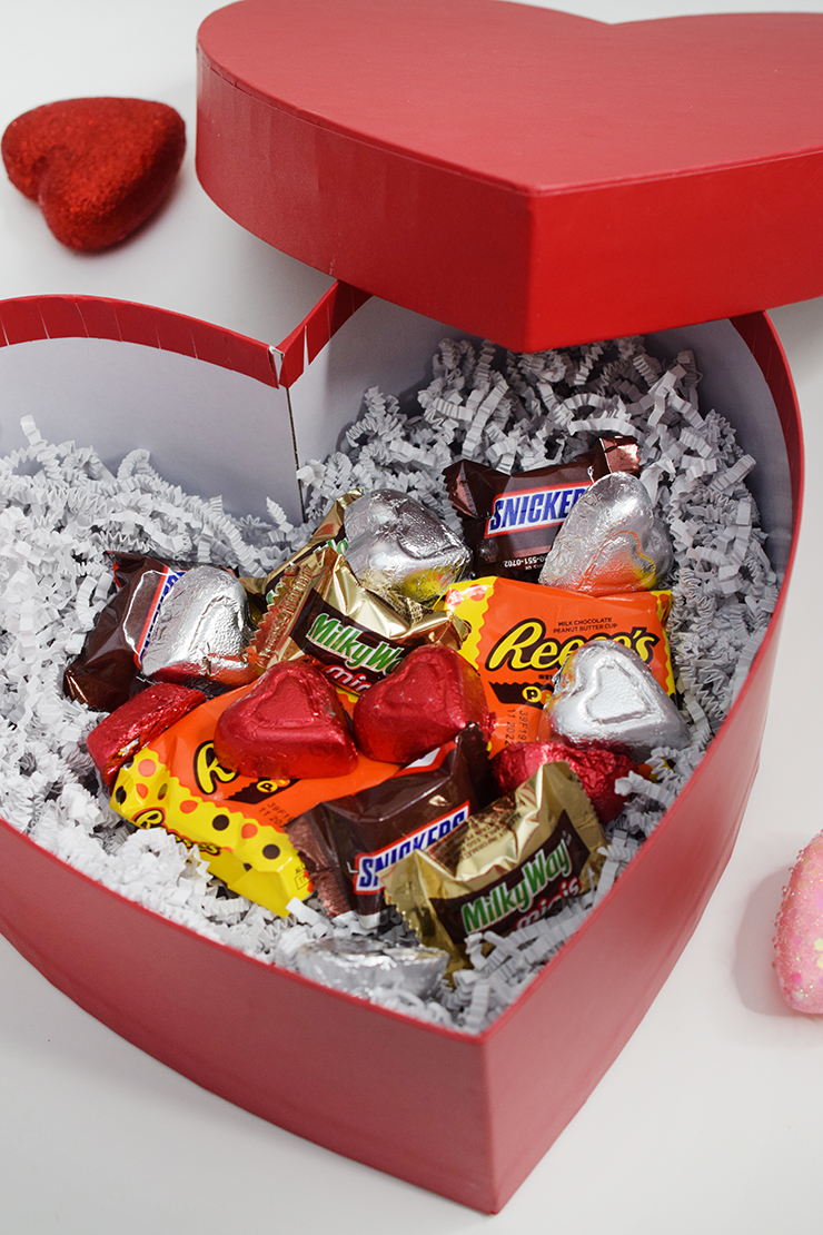 Red, heart-shaped box filled with crinkle paper and candy treats. 