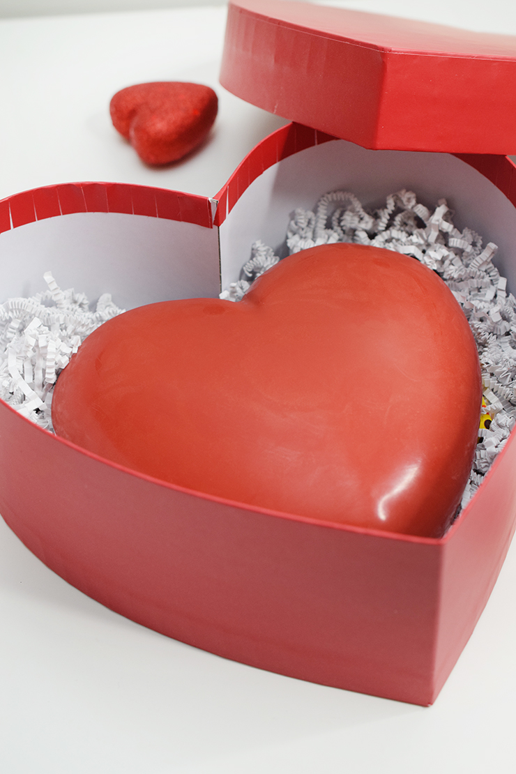 Red box shaped like a heart with a chocolate candy heart inside covering crinkle paper and candy.