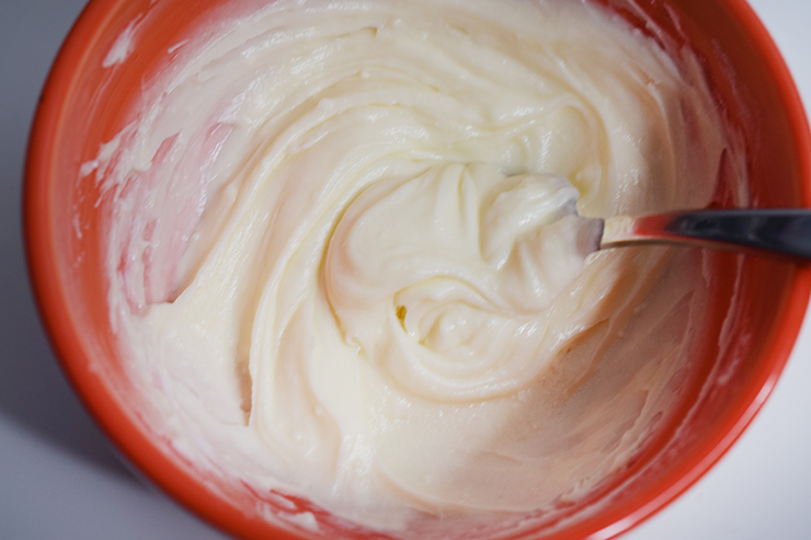 Homemade buttercream frosting in an orange bowl with a spoon to mix it together.