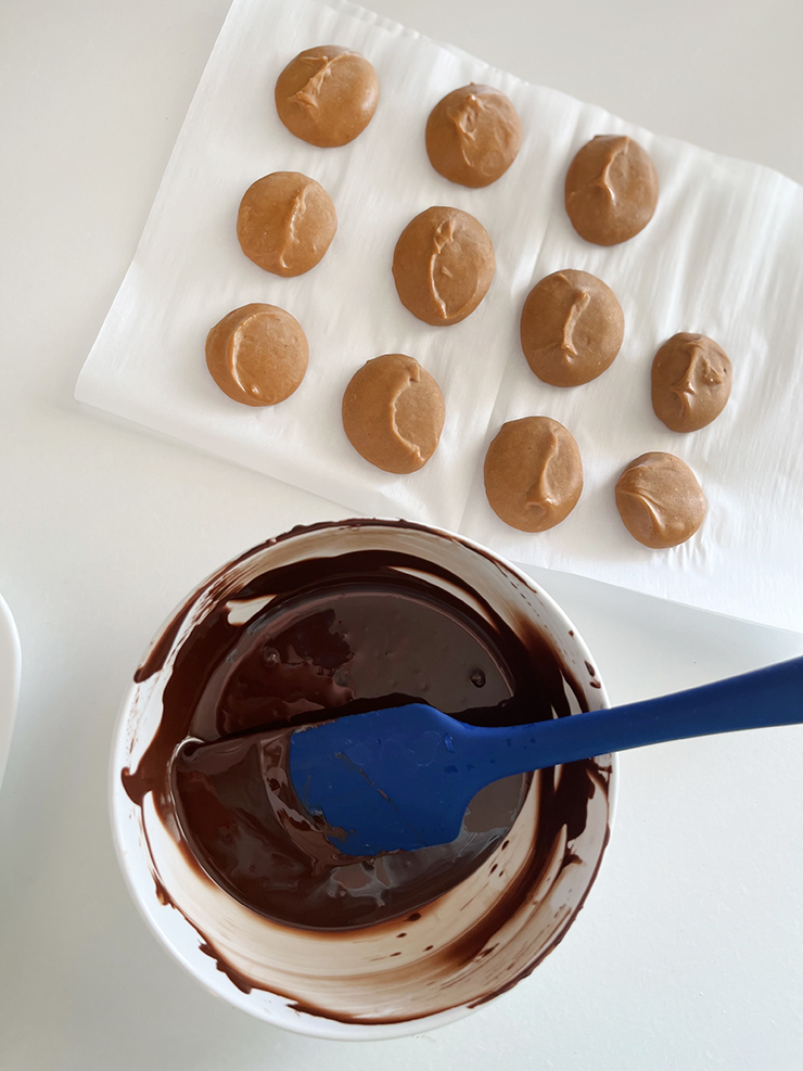 Parchment paper with chilled peanut butter spoonfuls sitting next to a bowl of melted chocolate.