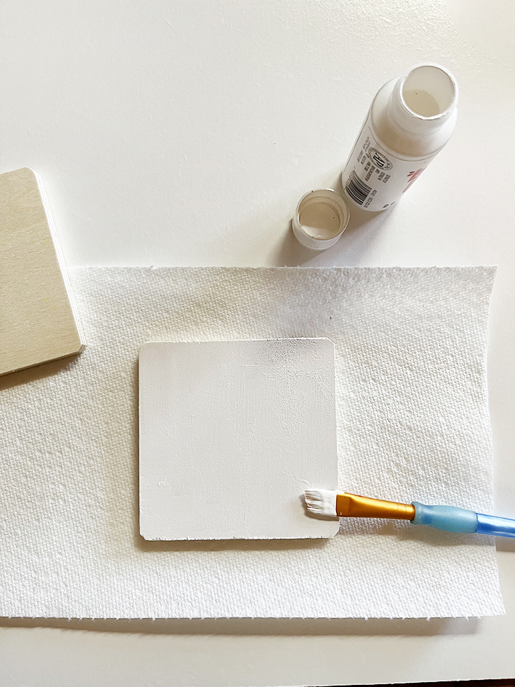 Wooden coaster painted white, sitting on a paper towel with a paint brush on top.