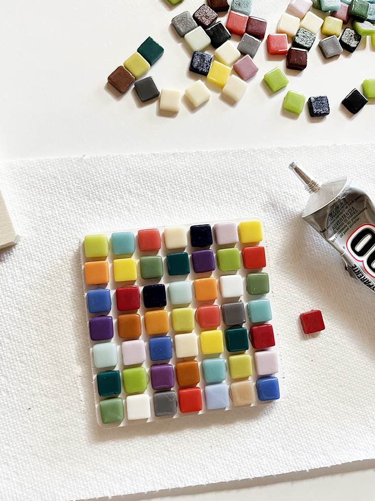 Small tiles on top of a square piece of wood with a container of glue beside it and more, loose tiles on the table.