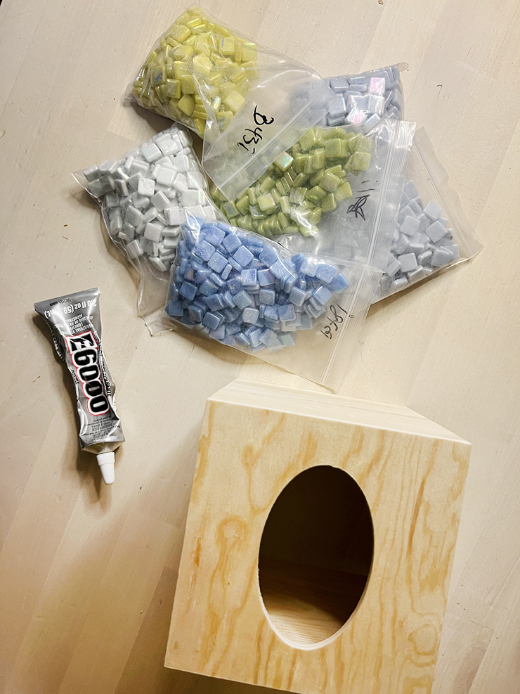 Materials to make a mosaic tile tissue box: wooden tissue box, E6000 glue, and baggies of small square tiles in shares of blue, green, and yellow.