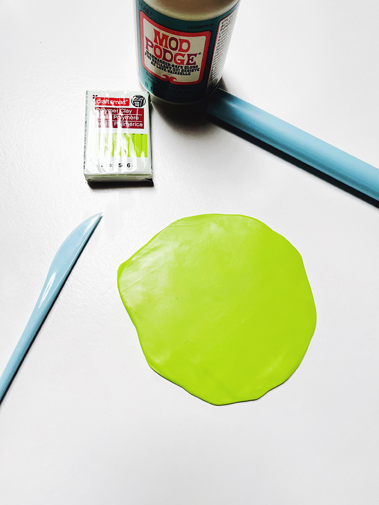 Green polymer clay rolled out on a white surface with clay tools and a bottle of mod podge in the background.