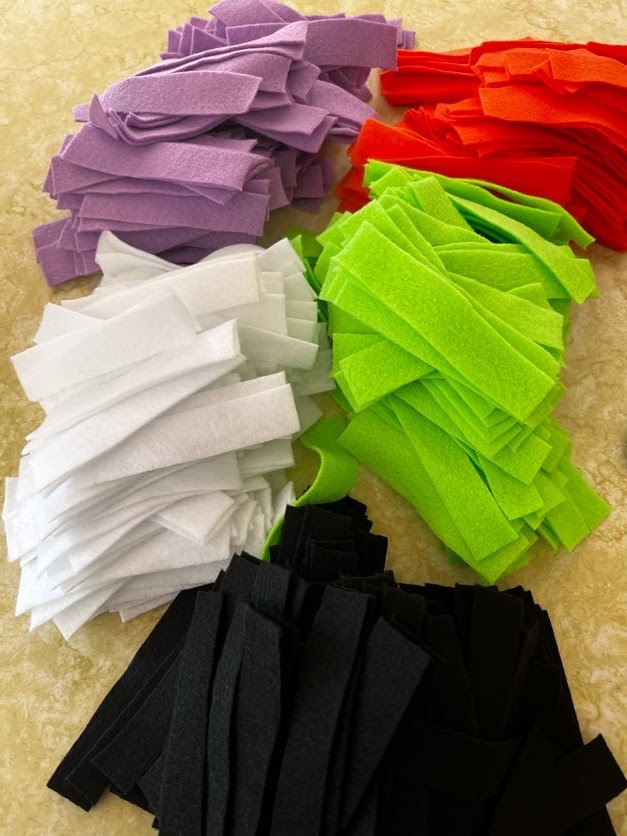 Five piles of felt in different colors (purple, orange, white, green, and black) cut into strips.