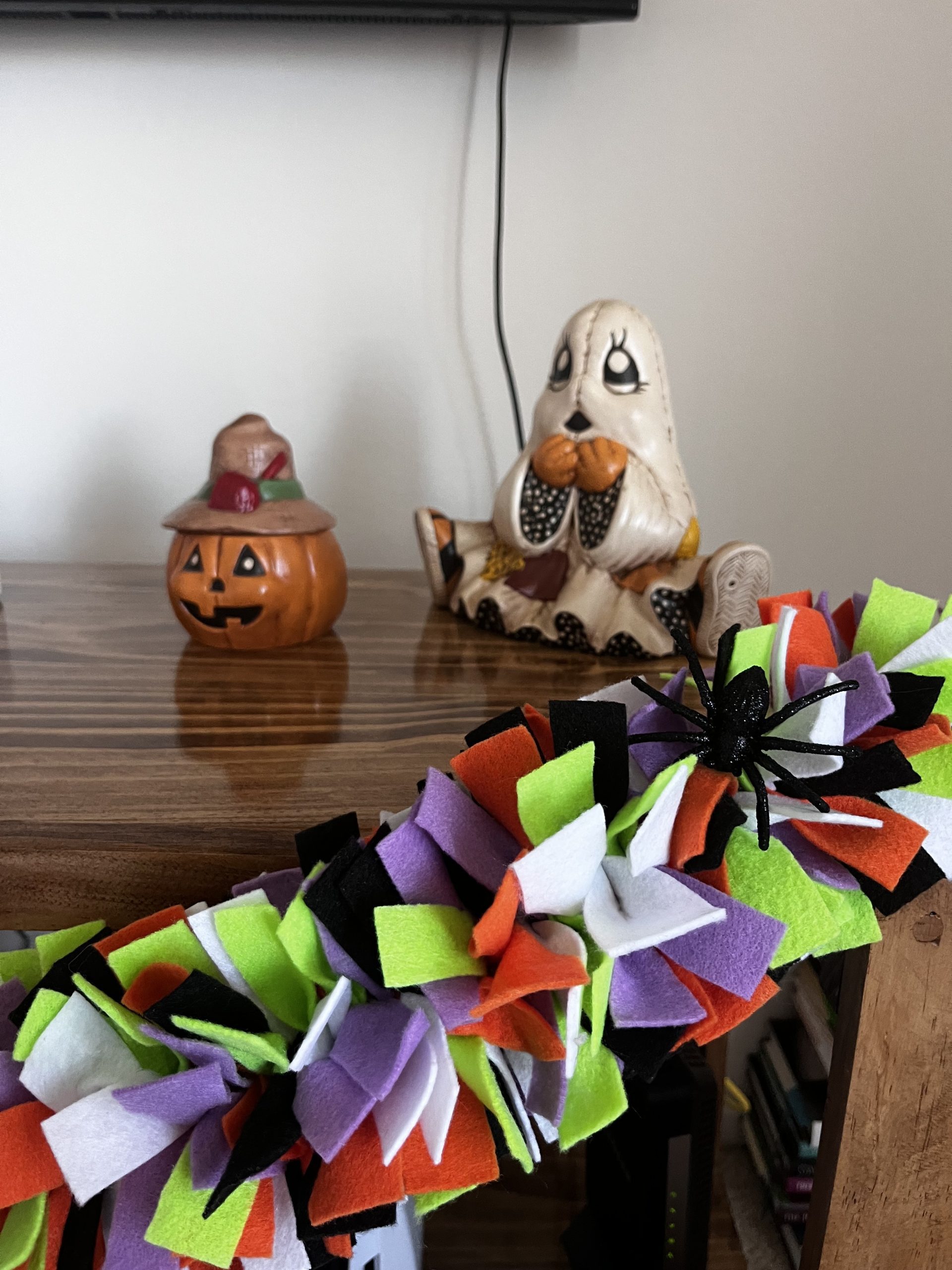 Multicolor felt garland attached to a wooden mantle with a fake spider sitting on the garland and two Halloween decorations behind. 
